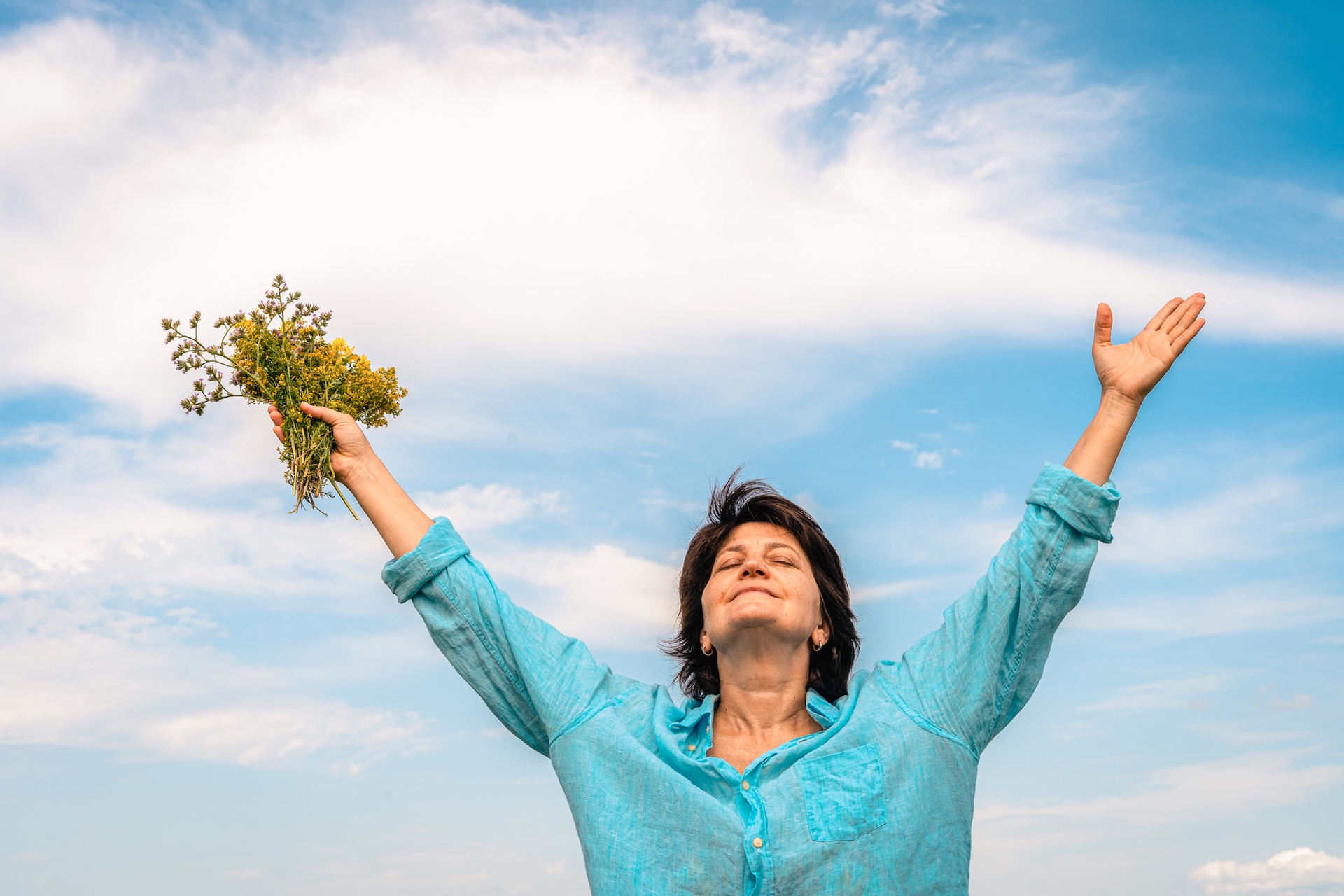 Woman standing in a green field with her arms outstretched, facing the sun, embracing the happiness and beauty of nature. Women's mental health, accepting one's age